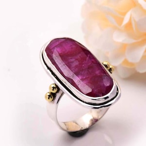 Può includere: Un anello d'argento con una grande pietra di rubino di forma ovale incastonata in una montatura d'argento. L'anello ha accenti dorati sui lati.