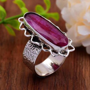May include: A silver ring featuring a large, faceted, oval-shaped ruby gemstone. The ruby is a deep, rich red color. The ring has an ornate, floral-inspired design on the band and around the gemstone setting. The ring is displayed on a wooden surface.