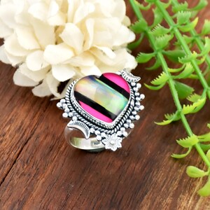 Op de afbeelding: Zilveren ring met een hartvormige steen. De steen heeft een roze, zwarte en iriserende vormgeving. De ring heeft een gedetailleerde, sierlijke zetting met kleine bloemen en halve maan accenten. Gepresenteerd op een houten oppervlak.
