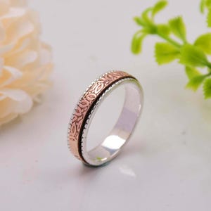 Puede incluir: Un anillo de plata con una banda de oro rosa que presenta un delicado diseño floral. El anillo es una pieza de joyería simple y elegante.