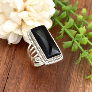 May include: A rectangular black onyx ring with a silver-colored band. The ring features a rope-like border around the onyx stone and a multi-band design. The ring is set against a wooden surface with white flowers and green foliage.