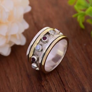 Könnte beinhalten: Ein drehbarer Ring aus Silber und Gold mit roten und weißen Edelsteinen. Der Ring hat eine strukturierte Band und ist so konzipiert, dass er sich frei drehen kann.