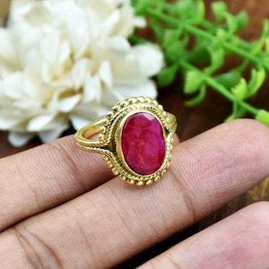 Puede incluir: Un anillo de tono dorado con una piedra preciosa de color rosa intenso en forma ovalada engastada en un bisel. La banda de metal y el engaste que rodea la piedra están decorados con un patrón texturizado de cuentas que crea un borde definido.