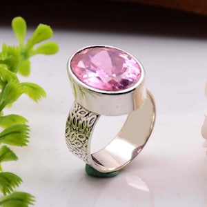 以下が含まれることがあります： 大きな楕円形のピンクの宝石が付いたシルバーリング。リングのバンドは、繰り返しのパターンでテクスチャ加工されています。