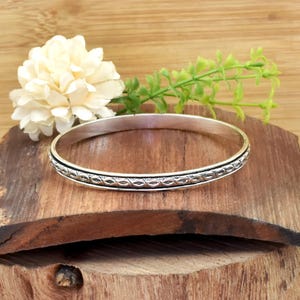 Puede incluir: Un brazalete de plata con un diseño grabado de formas ovaladas repetidas. El brazalete se muestra sobre una superficie de madera, con una flor blanca y ramitas verdes en el fondo.
