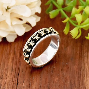 May include: A silver ring with a band of gold skulls. The skulls are raised and have a black outline. The ring is on a wooden surface.