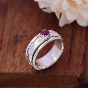 May include: A silver spinning ring with a red gemstone set in the center. The ring has a textured band and is designed to spin freely.