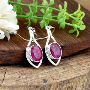 May include: A pair of silver drop earrings featuring oval-shaped, faceted deep pink ruby gemstones set in a polished metal frame. The earrings have a slender, elongated design with a hook closure, showcasing a vibrant contrast between the metallic silver and the dark red stones.