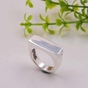 Puede incluir: Un anillo de plata con una banda rectangular. La banda es lisa y pulida en la parte superior y texturizada en los lados.