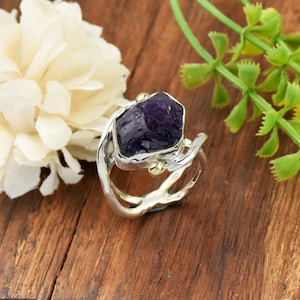 May include: A silver ring featuring a large, raw, dark purple amethyst gemstone. The ring has a unique, organic design with gold accents. The gemstone is set in a silver bezel, and the band has a flowing, textured appearance. The ring is displayed on a wooden surface.