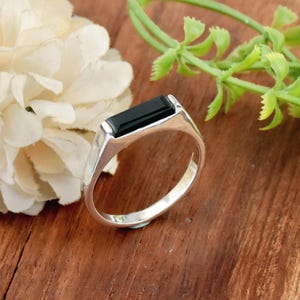 Könnte beinhalten: Ein silberner Ring mit einem schwarzen rechteckigen Edelstein. Der Ring hat ein schlichtes, klassisches Design.