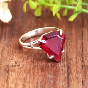 Pode incluir: Um anel de prata com uma grande gema vermelha facetada. A gema está engastada em garras e tem forma triangular.