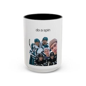Könnte beinhalten: Weißer Keramikbecher mit schwarzem Innenrand. Der Becher zeigt eine Gruppe von Hockeyspielern und den Satz "do a spin" in schwarzem Text. Die Hockeyspieler tragen grüne und schwarze Trikots.