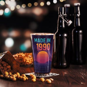 Op de afbeelding: Een helder pintglas met de tekst "MADE IN 1990" in blauw en oranje. Het glas heeft een retro zonsondergangontwerp met palmbomen en een rasterpatroon. Het glas staat op een onderzetter, met pinda's en flessen op de achtergrond.
