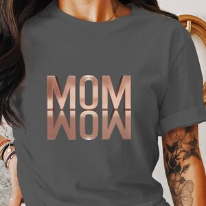 以下が含まれることがあります： 「MOM WOW」と書いてある、チャコールグレーのTシャツに、ローズゴールドのグラフィックが施されています。