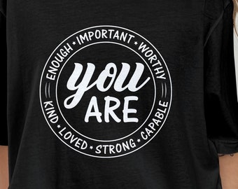 Op de afbeelding: Zwart T-shirt met een witte cirkelvormige afbeelding met de tekst "You Are" omringd door de woorden "Enough, Important, Worthy, Kind, Loved, Strong, Capable".