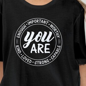 May include: Black t-shirt with a white circle graphic that says "You Are" surrounded by the words "Enough, Important, Worthy, Kind, Loved, Strong, Capable".