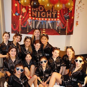 May include: A group of girls wearing black satin pyjamas with white trim and sunglasses pose in front of a red and gold movie night backdrop. The backdrop features a red curtain, a red carpet, and the words "Movie Night" in white lights.