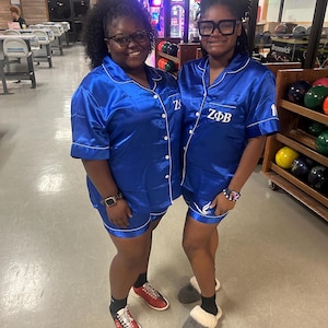 Könnte beinhalten: Zwei Personen tragen passende blaue Satin-Pyjama-Sets mit weißer Paspelierung. Die Hemden tragen die griechischen Buchstaben ZΦB. Eine Person trägt rot-schwarze Bowlingschuhe, die andere graue Hausschuhe.