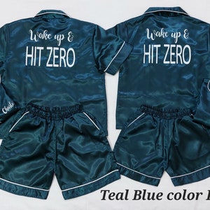 May include: Two teal blue satin pyjama sets with short sleeves and shorts. The tops have white embroidery that says "Wake up & HIT ZERO". One set has the name "Charle" embroidered on the shirt and the other set has the name "Sophi" embroidered on the shirt.