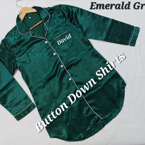 Peut inclure: Chemise boutonnée vert émeraude avec bordure blanche et le nom "David" brodé sur la poche poitrine. La chemise est en matière satinée brillante et est suspendue à un cintre en bois.