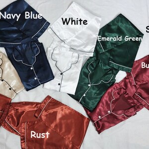 May include: Six pairs of satin pajamas in different colors: champagne, navy blue, white, emerald green, burgundy, and rust. Each set includes a shirt and pants. The text "PJ's Shirts with pants" is written on the image.