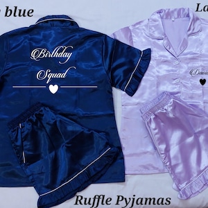 May include: Navy blue and lavender satin ruffle pajamas sets. The navy blue set has "Birthday Squad" printed on the back. The lavender set has "David" printed on the front. Both sets include a short-sleeved top and shorts.