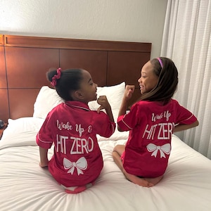 May include: Two girls wearing matching red satin pyjamas with white text that says "Wake Up & Hit Zero" and a white bow on the back. The girls are sitting on a white bed and smiling at each other.