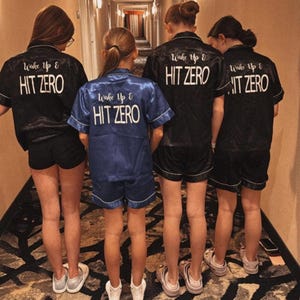 May include: Four people wearing black and blue satin pyjamas with the text "Wake Up & Hit Zero" on the back. The people are standing in a hallway.