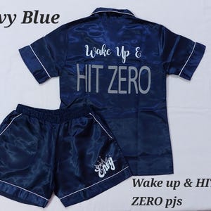 May include: Navy blue pyjama set with white trim. The short-sleeved top has the text "Wake Up & HIT ZERO" on the back. The shorts have the name "Envy" printed on the leg. The set is made of a shiny, satin-like material.