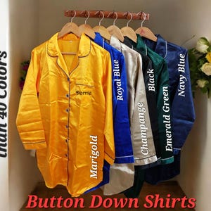 Può includere: Camicie button-down in vari colori, tra cui giallo calendula, blu reale, nero, verde smeraldo e blu navy. Le camicie sono esposte su grucce di legno. Il testo sull'immagine dice "Disponibile in più di 40 colori."