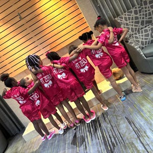 May include: Seven girls wearing pink satin pyjamas with white text that says "Level Up" and "Hit Zero". The girls are standing in a line, facing the camera.