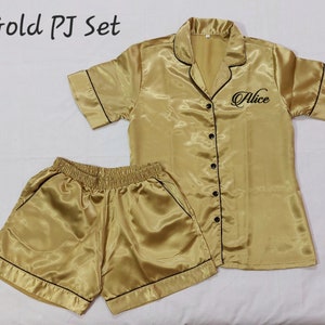 May include: A gold satin pyjama set with black piping. The set comprises a short-sleeved button-down shirt and matching shorts. The shirt is embroidered with the name "Alice". The image also includes the text "Gold PJ Set".