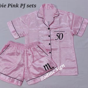 May include: Pink satin pajama set with black trim. The short-sleeved top has a collar and the number 50 printed on the front. The shorts have an elastic waistband and the letter "m" printed on the leg. The text "Barbie Pink PJ sets" and "Zodiac Sign" are also visible.