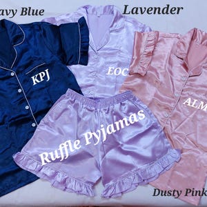 May include: A collection of satin pajama sets in navy blue, lavender, and dusty pink. Each set includes a button-down shirt and shorts. The lavender shorts have a ruffle trim and the text "Ruffle Pyjamas". The shirts have initials embroidered on them.