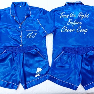 May include: A blue satin pyjama set with white trim. The top has a button-up front and the text "Twas the Night Before Cheer Comp" on the back. The shorts have a white "Liberty" logo on the right leg. The top has the initials "ELJ" embroidered on the left chest.