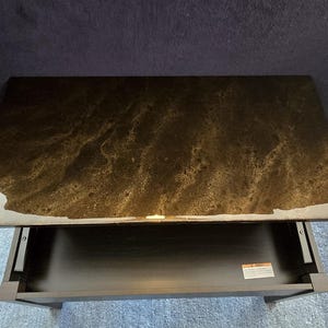 May include: A dark brown end table with a drawer and a glossy epoxy resin top. The top has a marbled effect with swirls of gold and brown.