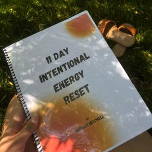 May include: A white spiral-bound book with the title 11 DAY INTENTIONAL ENERGY RESET and the author name MORGAN MITCHELL printed in black text on the front cover, featuring soft orange and red circular abstract shapes across the surface.
