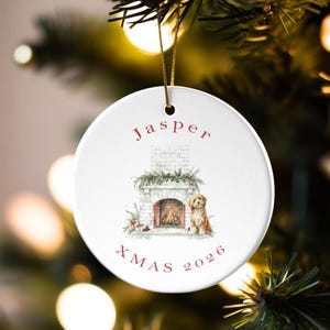 Puede incluir: Un adorno navideño redondo de cerámica blanca con una ilustración en acuarela de una chimenea de ladrillo con vegetación y un perro marrón dorado sentado al lado. El texto Jasper está impreso arriba y XMAS 2026 debajo.