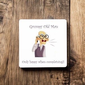 Op de afbeelding: Witte vierkante onderzetter met een cartoonillustratie van een oude man met een bril die aan het bellen is. Tekst luidt "Grumpy Old Man" en "Only happy when complaining!!". De onderzetter ligt op hout.