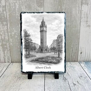 May include: A black and white print of the Albert Clock on a slate plaque. The detailed illustration shows the clock tower in a city square, with the text "Albert Clock" below the image. The plaque is displayed on a black stand.