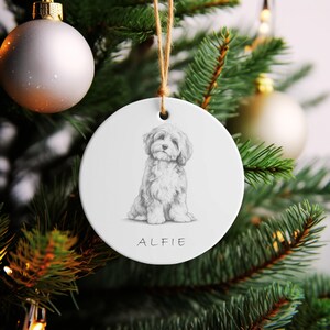 Könnte beinhalten: Ein runder, weißer Keramik-Weihnachtsbaumschmuck mit einer detaillierten schwarz-weißen Bleistiftzeichnung eines flauschigen Hundes in sitzender Position. Unter der Illustration steht der Name ALFIE in einfachen schwarzen Großbuchstaben. Er hängt an einer braunen Schnur.