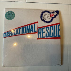 May include: A white album cover with the words "INTERNATIONAL RESCUE" in blue and red lettering. The album cover also features a logo with a hand and the letters "IR". A small sticker is in the upper left corner.