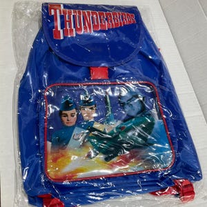 May include: Blue backpack with a clear front pocket displaying a graphic of Thunderbirds characters. The backpack has red straps and a red clasp. The word "THUNDERBIRDS" is printed across the top in red and white.