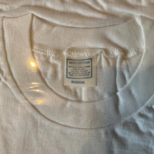 May include: Close-up of a white, crew neck t-shirt. The shirt has a tag that reads "100% COTTON" and "MEDIUM". The fabric appears to be slightly textured. The shirt is likely vintage or a classic style.