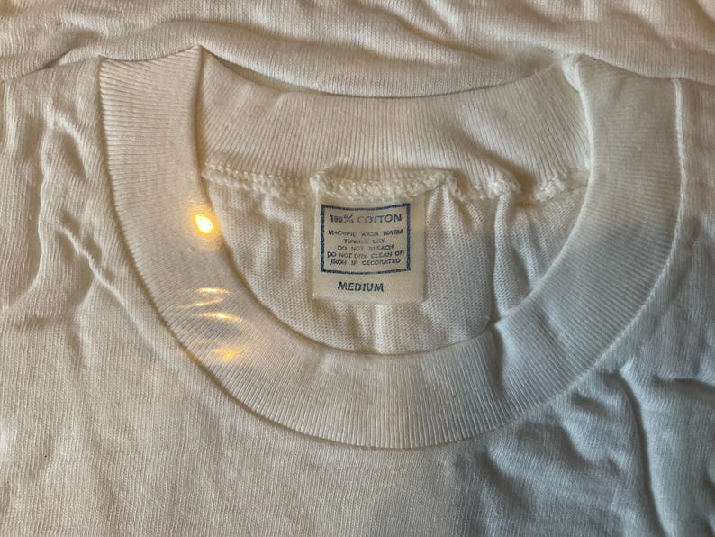 May include: Close-up of a white, crew neck t-shirt. The shirt has a tag that reads "100% COTTON" and "MEDIUM". The fabric appears to be slightly textured. The shirt is likely vintage or a classic style.