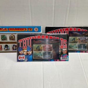 May include: Three sets of collectible postage stamps featuring Classic Children's TV and Thunderbirds themes. The packaging displays colourful character illustrations, the Thunderbirds logo, and individual stamp designs encased in clear plastic sleeves.