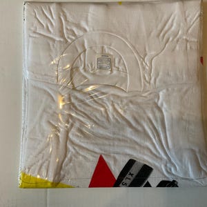 May include: A white t-shirt, folded and sealed in clear plastic. The shirt has a crew neck and a tag visible at the neckline. The bottom corner of the shirt shows a red triangle and black and gray stripes with the text "XLS".