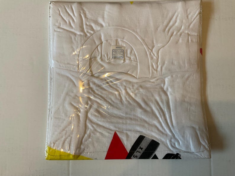 May include: A white t-shirt, folded and sealed in clear plastic. The shirt has a crew neck and a tag visible at the neckline. The bottom corner of the shirt shows a red triangle and black and gray stripes with the text "XLS".