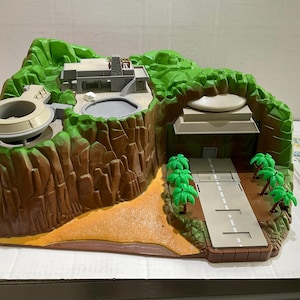 Puede incluir: Un set de juego de plástico que presenta un paisaje de montaña rocosa marrón con colinas verdes, una estructura de edificio gris, una piscina circular y una pista de aterrizaje con pequeñas palmeras verdes. Incluye una hoja de pegatinas al lado.